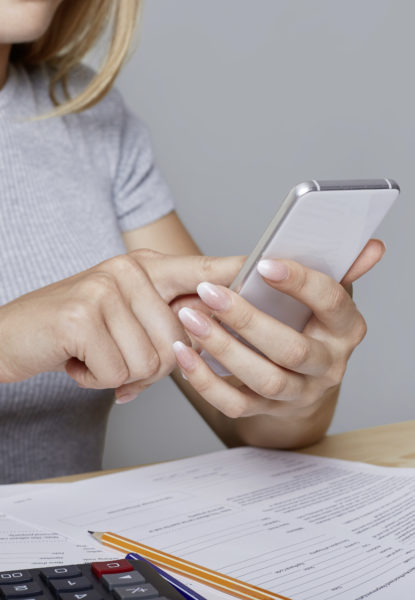 Unrecognizable woman holds modern mobile phone in hands, sits at work table, surrounded with documents, laptop, pencil, calculator, downloads necessary information from internet. Job and technology Downloads